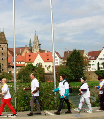Personen laufen mit Walkingstöcken am Neu-Ulmer Donauufer, im Hintergrund die Stadtsilhouette von Ulm 