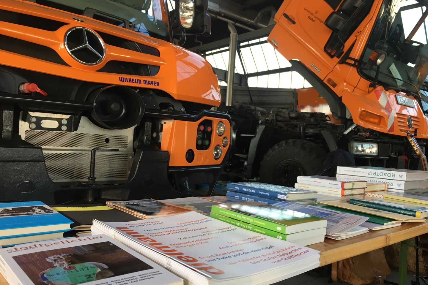 Ein Tisch mit Bücherstapeln, direkt dahinter parken zwei große Nutzfahrzeuge 