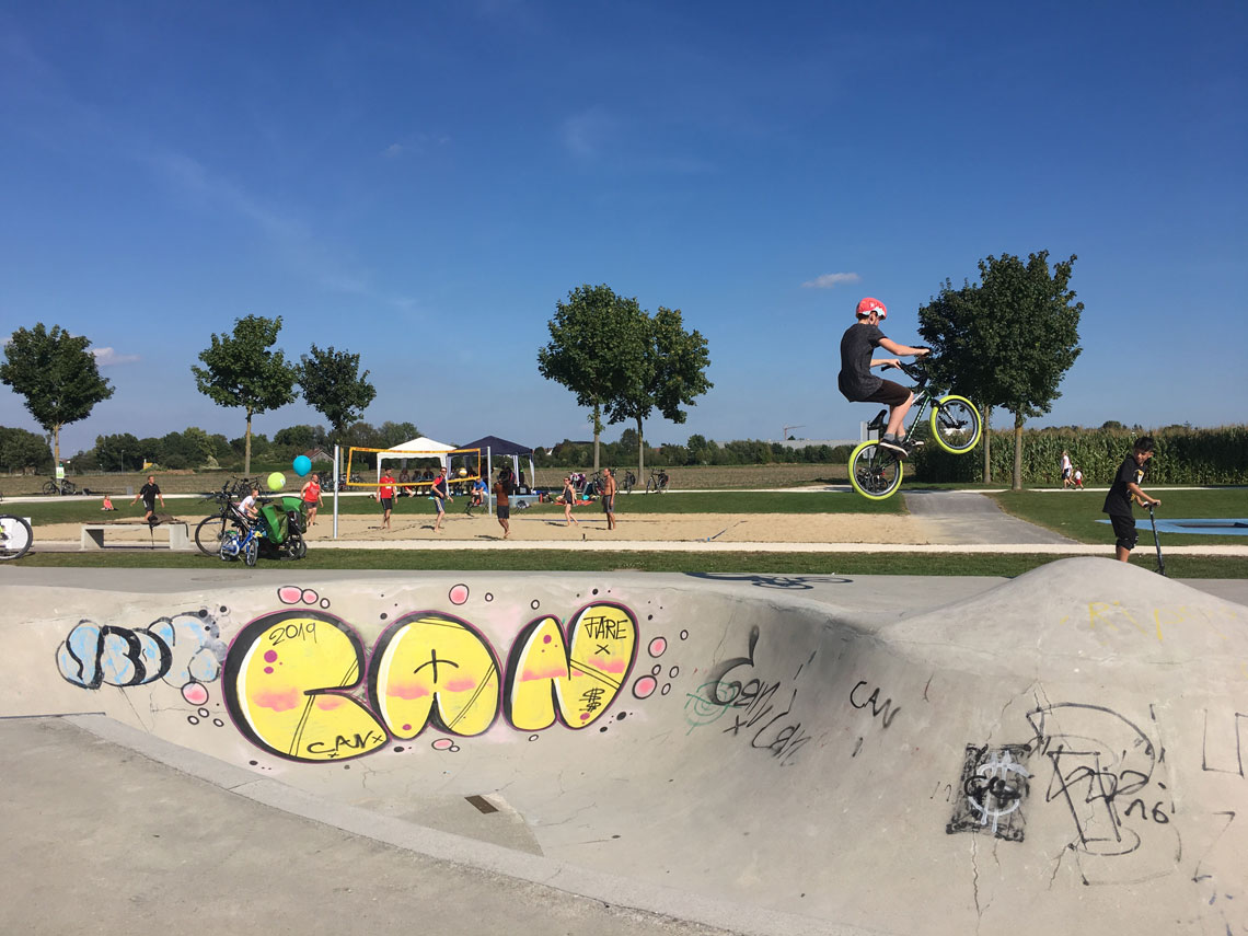 Ein BMX-Radfahrer springt mit seinem Bike im Skatepool im Sport- und Freizeitpark Wiley in die Luft, im Hintergrund spielen Personen Beachvolleyball