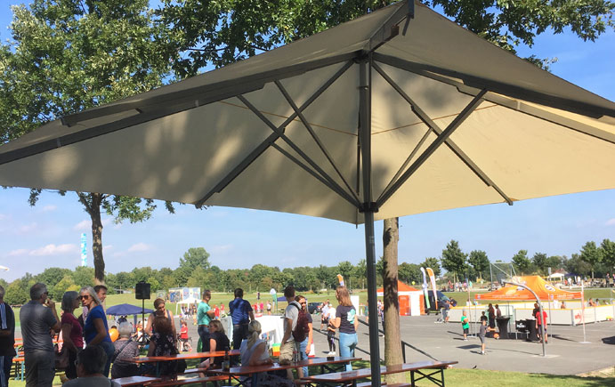 Besucher der Veranstaltung "Sport trifft Trend" sitzen unter einem Sonnenschirm an Biertischen, im Hintergrund Sportstände und Besucher