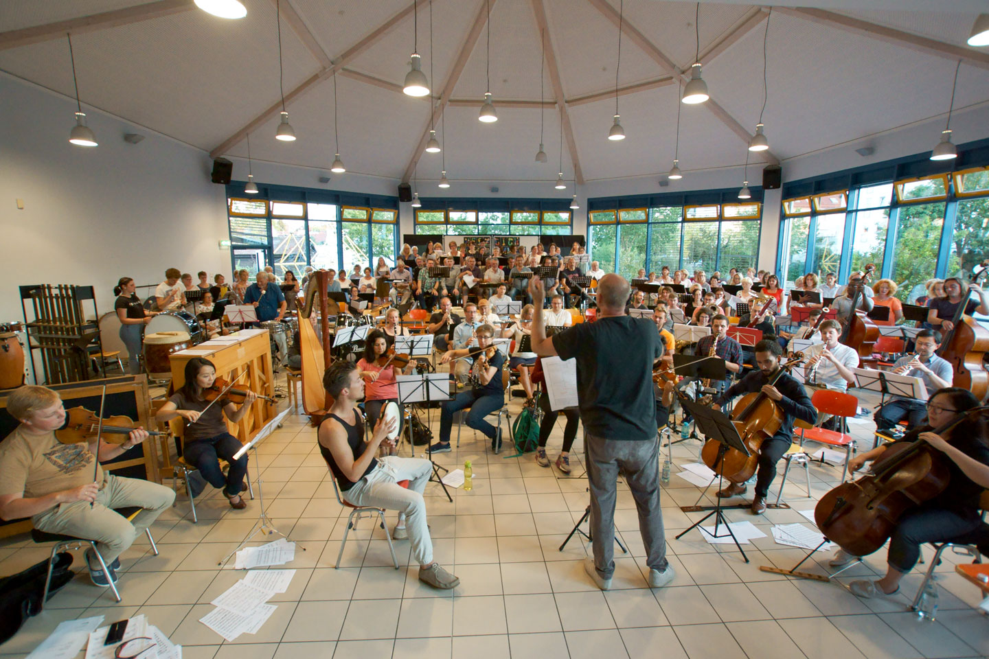 Ein Saaler voller Musiker mit verschiedenen Instrumenten bei einer Probe, im Vordergrund der Dirigent 