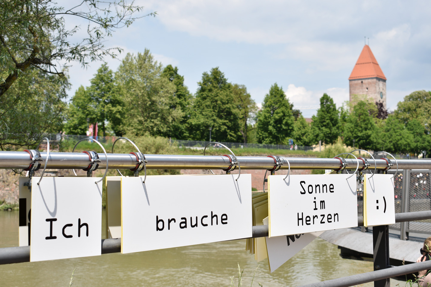 Kunstinstallation am Neu-Ulmer Donauufer mit Klapptafeln, die den Satz "Ich brauche Sonne im Herzen" bilden, im Hintergrund der Ulmer Gänsturm 