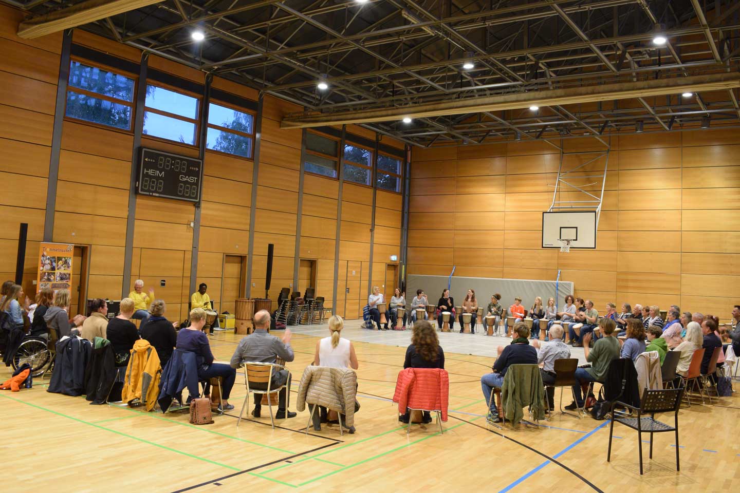 Ca. 40 Personen sitzen in einem großen Stuhlkreis in einer Turnhalle und trommeln 