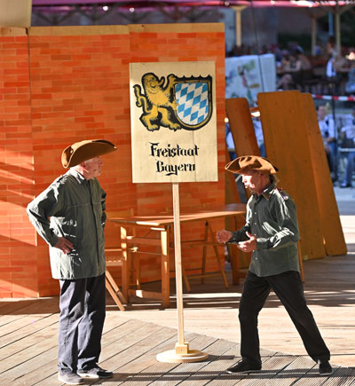Foto von der Aufführung des Bürgertheaters "Napoleon geht baden": Zwei Schauspieler stehen auf der Bühne, zwischen ihnen ein Schild mit der Aufschrift "Freistaat Bayern" 