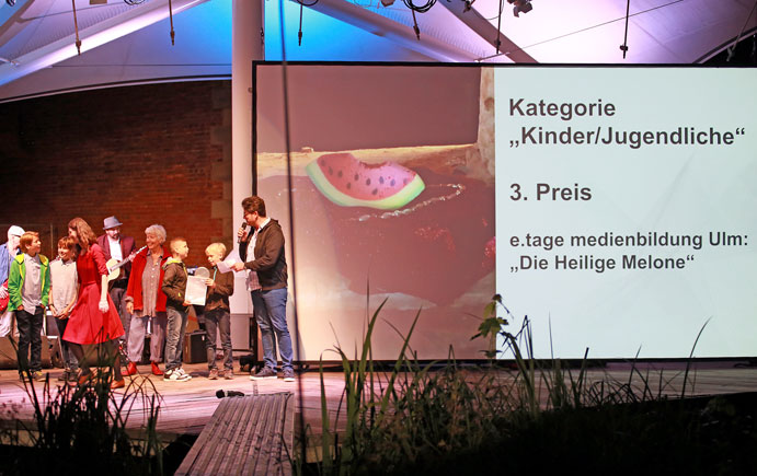 Auszeichnung der Gewinner beim Kurzfilmfestival auf der Bühne, rechts auf der Leinweind der Text "Kategorie Kinder/Jugenliche, 3. Preis: e.tage medienbildung Ulm: Die Heilige Melone"