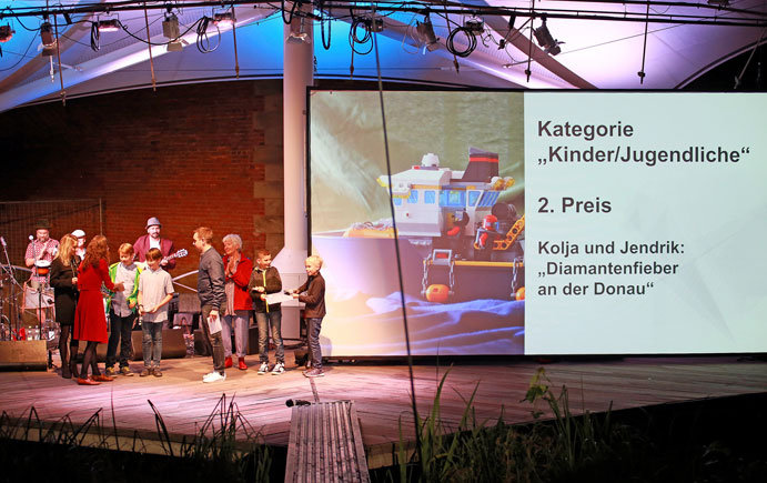 Auszeichnung der Gewinner beim Kurzfilmfestival auf der Bühne, rechts auf der Leinweind der Text "Kategorie Kinder/Jugenliche, 2. Preis: Kolja und Jendrik: Diamantenfieber an der Donau"