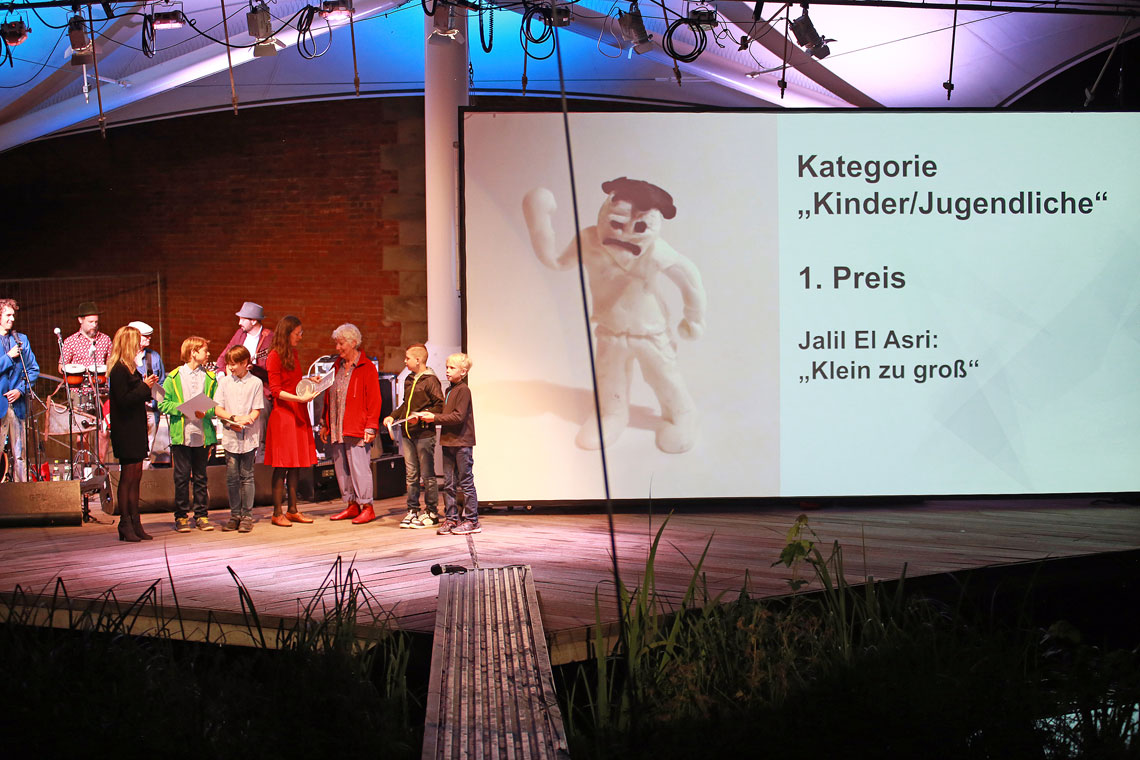 Auszeichnung der Gewinner beim Kurzfilmfestival auf der Bühne, rechts auf der Leinweind der Text "Kategorie Kinder/Jugenliche, 1. Preis: Jalil El Asri: Klein zu groß"