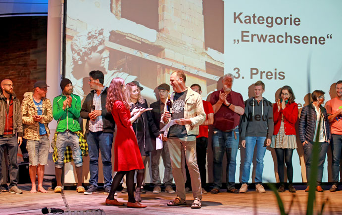 Die Moderatorin übergibt einem der Gewinner auf der Bühne einen Preis, im Hintergrund die anderen Filmemacher, an der Leinwand der Text "Kategorie Erwachsene, 3. Preis"