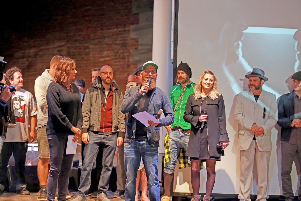 Einer der Gewinner des Kurzfilmfestivals spricht auf der Bühne zum Publikum, dahinter stehend die Moderatoren und weiteren Filmemacher
